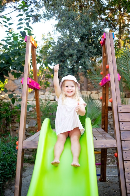 Un niño niña alegre se desliza por un tobogán en el patio de recreo y se ríe en el verano con un sombrero de Panamá y los pies descalzos