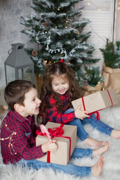 Niño y niña con camisas rojas sostienen cajas de regalo cerca del árbol de Navidad en el interior de Navidad