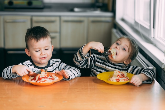 Niño y niña en la cocina comiendo pasta