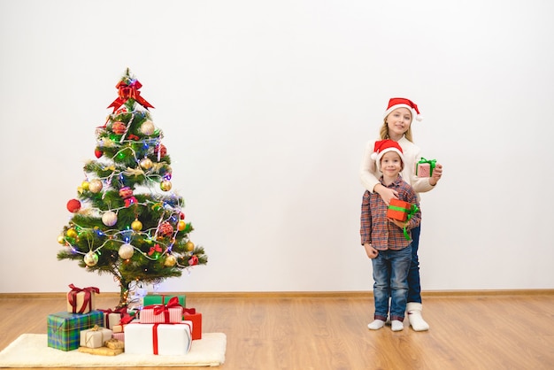 El niño y una niña están parados con cajas de regalo cerca del árbol de navidad.