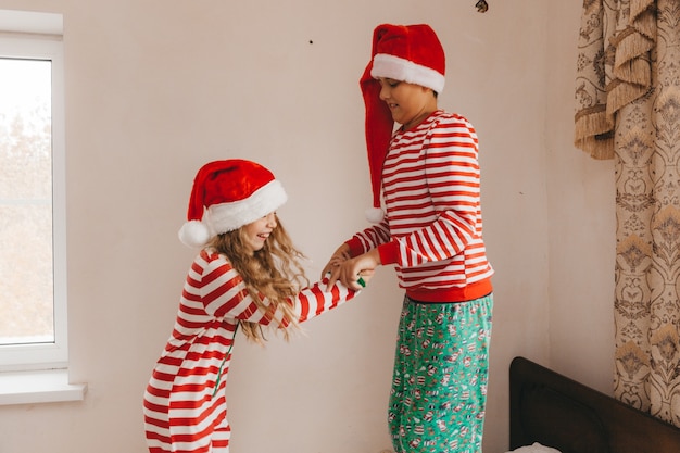 Niño y niña felices con sombreros de Navidad en la mañana de Navidad juegan y luchan con almohadas en la cama