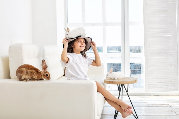 Niño niña hermosa y feliz sentada en el sofá con pequeños animales conejo en un sombrero