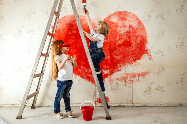 Un niño y una niña en jeans y una camiseta blanca se miran, con un pincel y un cubo en una escalera pintan la pared en rojo, un gran corazón.