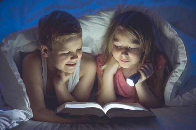 El niño y la niña con un libro y una linterna yacían a cubierto. Noche