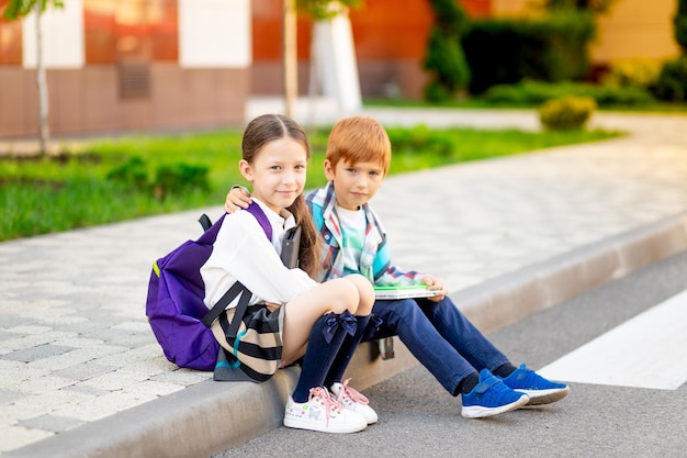 Un niño y una niña con maletines o mochilas están sentados en la escuela y leyendo un libro y una tableta volviendo a la escuela hermano y hermana están estudiando haciendo la tarea en la calle