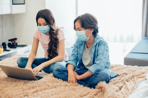 Niño y niña con máscaras médicas mientras permanecen en cuarentena y se sientan en la cama con una computadora portátil moderna