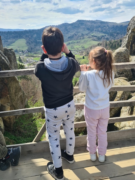 Un niño y una niña se paran en una cornisa, mirando las montañas.