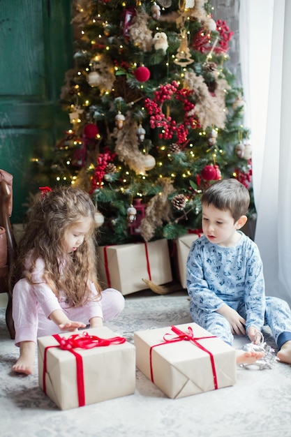 Niño y niña en pijama sostienen cajas de regalo cerca del árbol de Navidad en el interior de Navidad