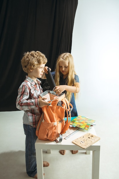 Niño y niña preparándose para la escuela después de un largo receso de verano. De vuelta a la escuela. Pequeños modelos caucásicos empacando una bolsa juntos sobre fondo de estudio. Concepto de infancia, educación, vacaciones o deberes.