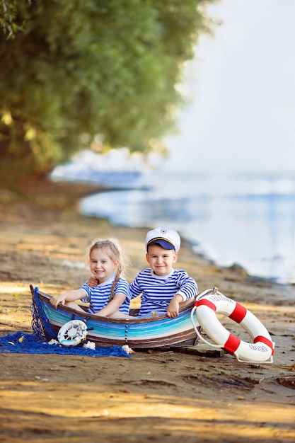 Foto niño y niña sentada en un barco, vestido como marinero, en una playa de arena con conchas junto al mar