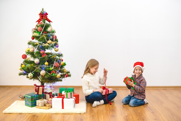 El niño y una niña se sientan con cajas de regalo cerca del árbol de navidad.