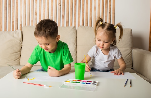 Un niño y una niña se sientan en una mesa y dibujan con pinturas sobre papel