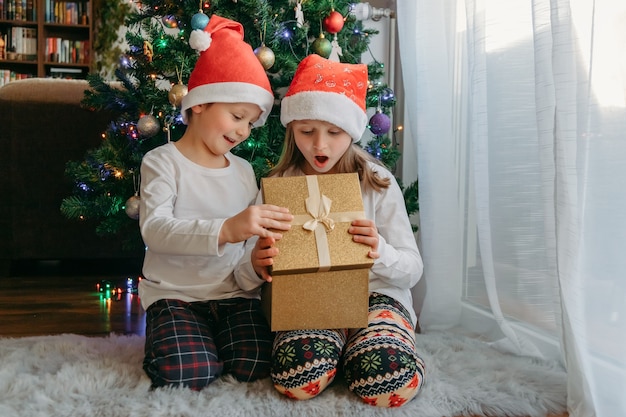 Foto un niño y una niña con sombreros navideños abren una caja de regalo debajo de un árbol de navidad. celebración, recepción de regalos, alegría de los niños. milagro, presente.