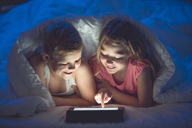 El niño y la niña con una tableta y una linterna yacían debajo del edredón. Noche