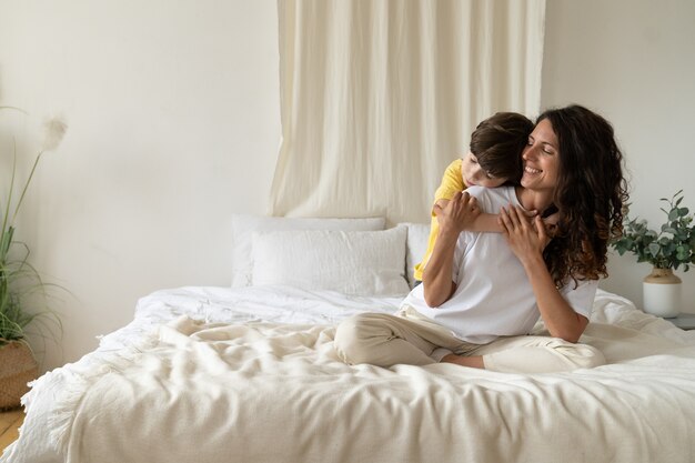 Niño, niño, abrazar, sonriente, mamá, en, mañana, sentarse, en, cama, llevando, pijama, en casa, el, fin de semana, amor familiar