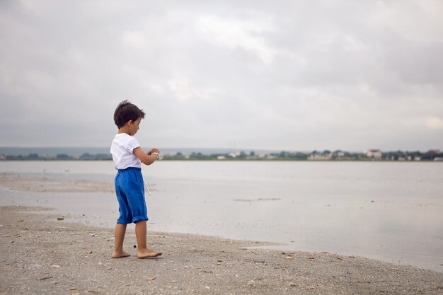Niño niño bronceado en pantalones cortos azules y una camiseta blanca camina descalzo en el lago en verano