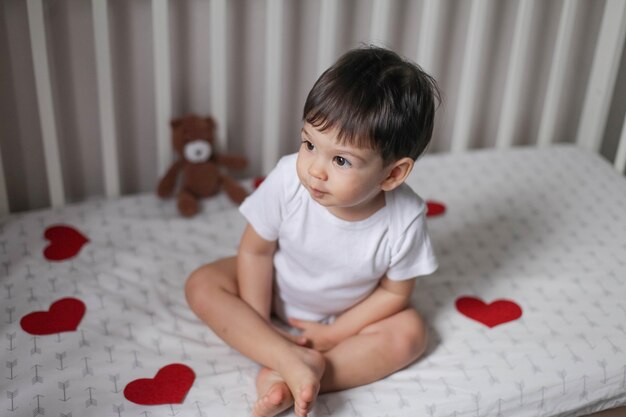Foto niño niño con cabello oscuro en un cuerpo blanco sentado en la cama blanca debajo de guirnaldas de corazones rojos en el día de san valentín y sonriendo.