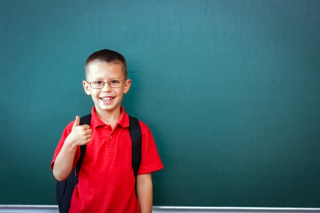 Un niño niño feliz de pie en la pizarra con una mochila escolar con gafas