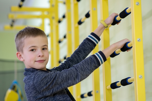 Niño niño haciendo ejercicio en una barra de escalera de pared dentro de la sala de gimnasia deportiva en una escuela.