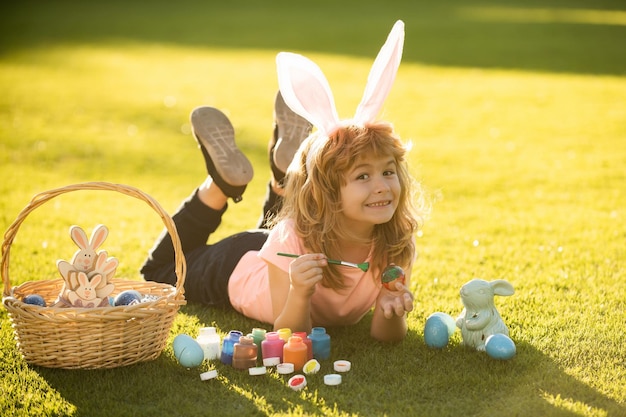 Niño niño con huevos de Pascua y orejas de conejo sobre hierba pintando huevos