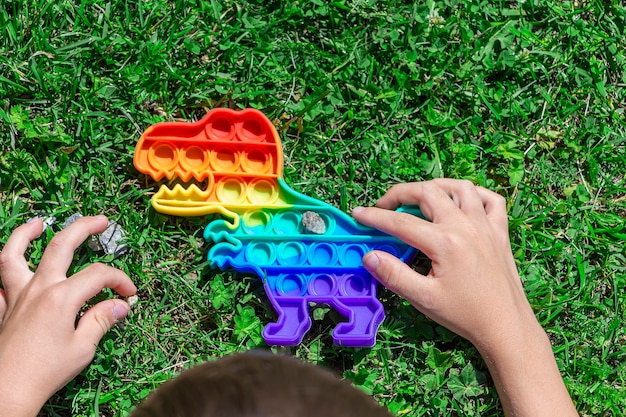 Niño niño juega con pop sobre el césped en el parque. Nuevo juguete de desarrollo sensorial en forma de dinosaurio con coloridas burbujas de silicona de arcoíris. Juego de inquietud de moda, antiestrés y alivio de la ansiedad.