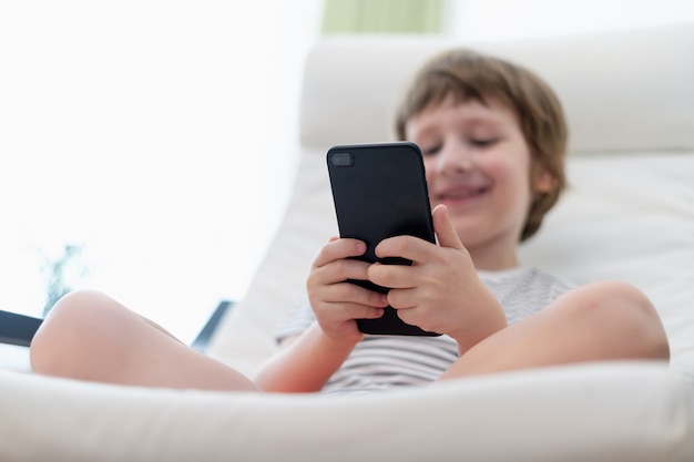 Niño niño jugando juegos en línea viendo videos en el teléfono inteligente sentado en el sofá entretenido en