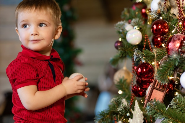 Foto niño niño mantenga la bola en el árbol de navidad. el niño espera vacaciones y regalo para año nuevo