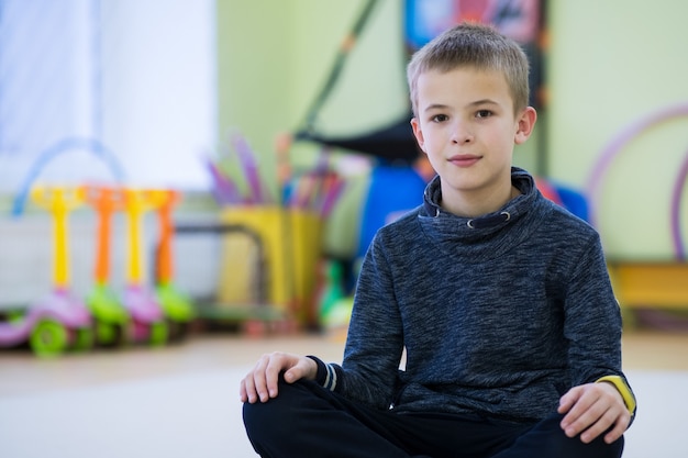 Niño niño sentado y relaxiong en el piso dentro de la sala de deportes en una escuela después del entrenamiento.