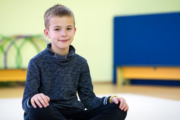 Niño niño sentado y relaxiong en el piso dentro de la sala de deportes en una escuela después del entrenamiento.