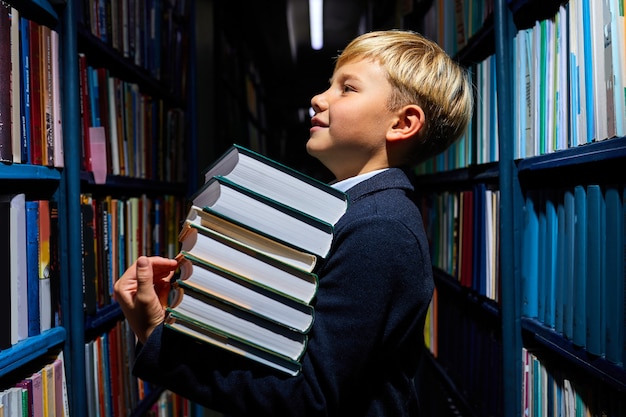 niño niño sosteniendo una pila de libros en la biblioteca de la escuela, preparándose para la educación escolar, pararse entre los estantes