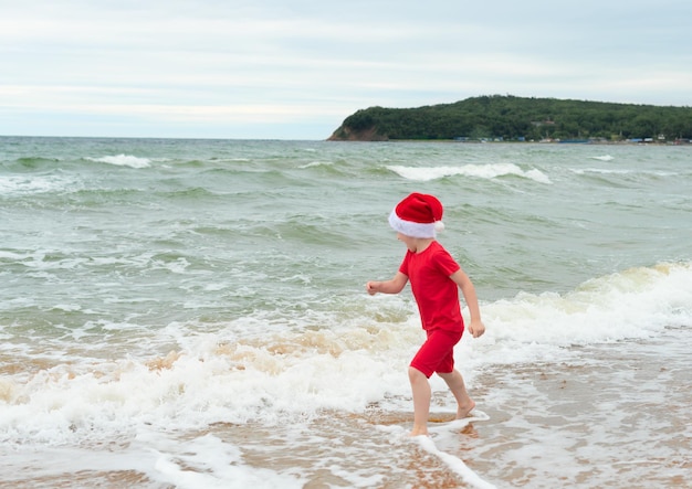 Niño niño en tela roja y sombrero de Santa Claus disfrutando de la Navidad en la playa en vacaciones de Navidad concepto de celebración de Navidad o Año Nuevo