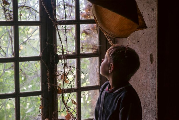 Un niño de nueve años cerca de una ventana de madera muy antigua cubierta de hiedra en una casa abandonada
