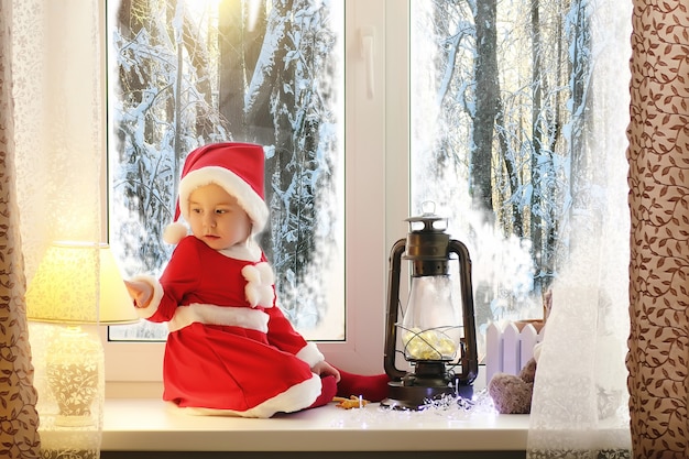 Un niño en el nuevo año mira por la ventana. Los niños esperan a Santa Claus. Un niño con traje de Santa se sienta en la ventana.