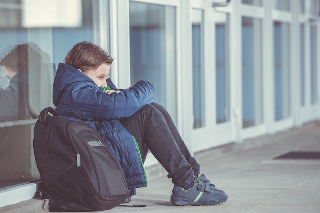 Foto niño o niño sentado solo en el piso frente a la escuela después de sufrir un acto de acoso escolar