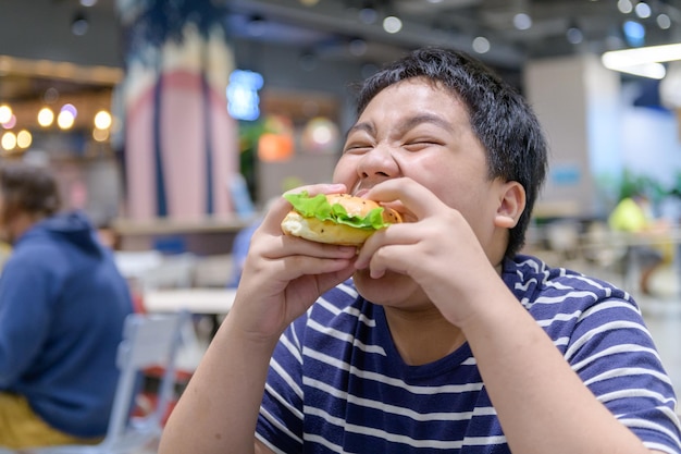 Un niño obeso come una hamburguesa en un patio de comidas en un centro comercial de comida chatarra
