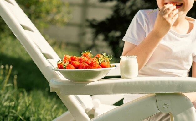 Un niño de ocho años come fresas con yogur en verano en el patio.