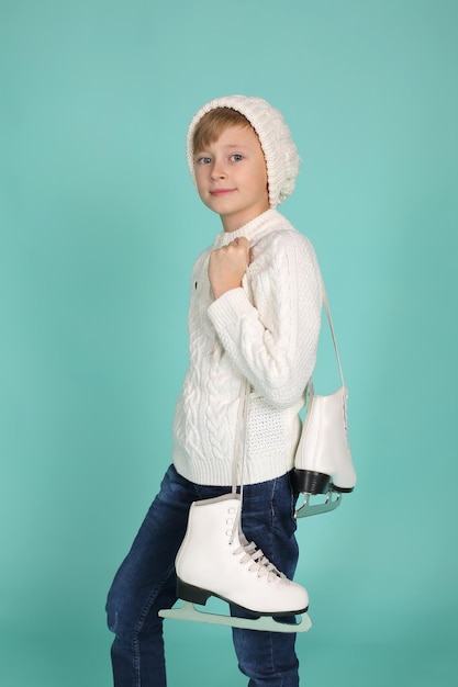 niño de ojos azules con un suéter de punto blanco y un sombrero de invierno sostiene patines de patinaje artístico en sus manos