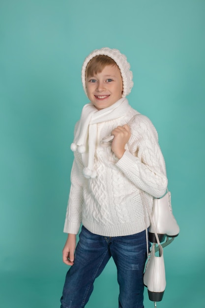 niño de ojos azules con un suéter de punto blanco y un sombrero de invierno sostiene patines de patinaje artístico en sus manos