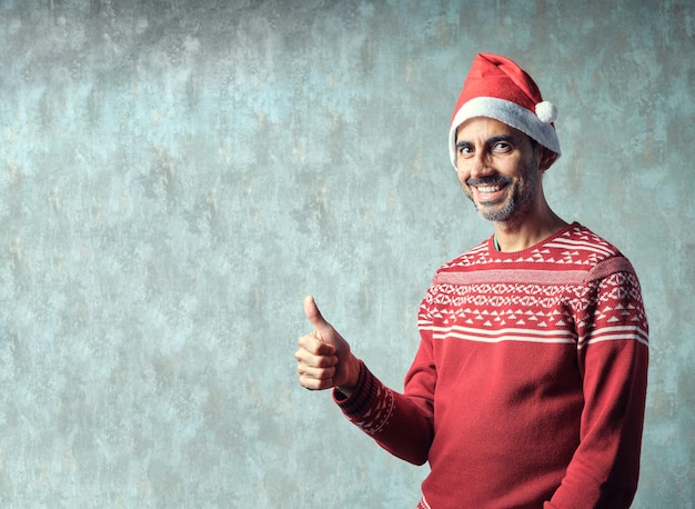 Niño oscuro con barba gris corta sonriendo en suéter rojo de Navidad y sombrero de Navidad con el pulgar hacia arriba