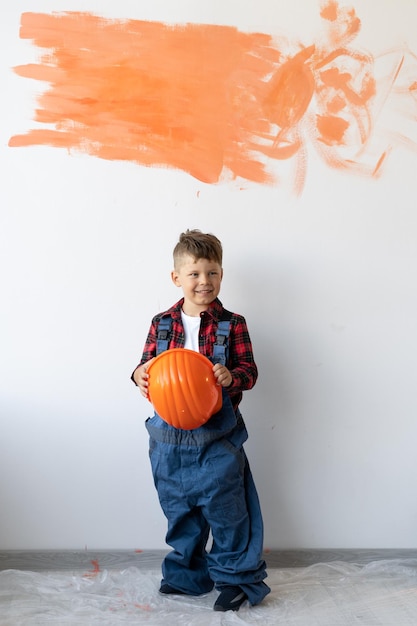 Foto un niño en overol sostiene un casco de seguridad en sus manos contra el fondo de un muro de hormigón blanco con una mancha naranja para el texto renovación de la casa o apartamento el niño ayuda a los padres a pintar