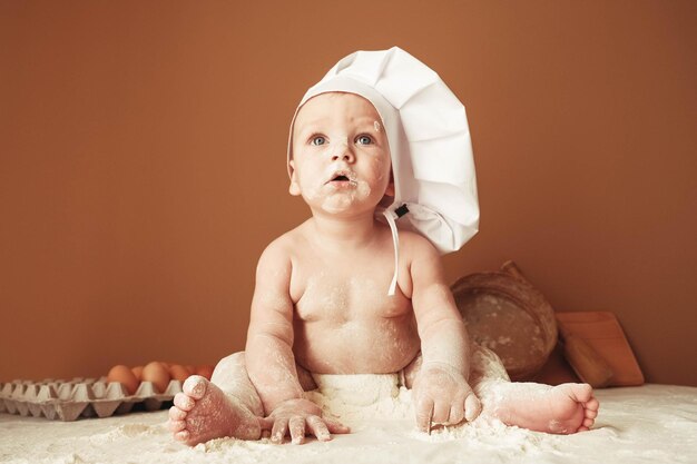 Niño panadero con sombrero de chef sentado en la mesa jugando con harina en un fondo marrón