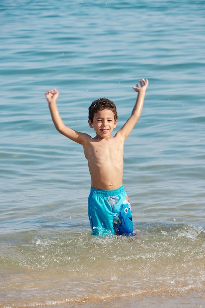 Un niño con pantalones cortos azules se para en el agua con los brazos en el aire.