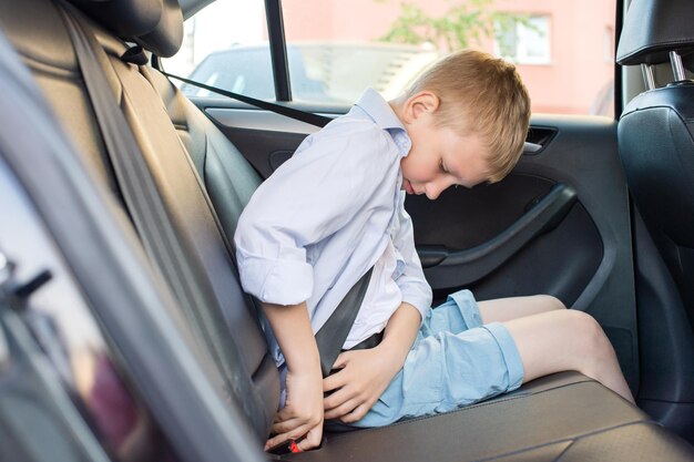 Un niño en pantalones cortos y una camisa está sentado en el coche y abriendo su cinturón de seguridad