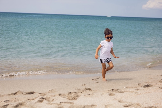 Niño en pantalones cortos a rayas y una camiseta blanca camina en una playa de arena y gafas de sol