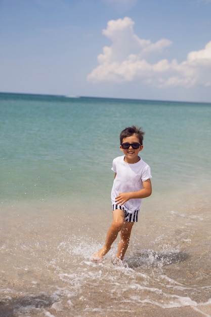 Niño en pantalones cortos a rayas y una camiseta blanca camina en una playa de arena y gafas de sol