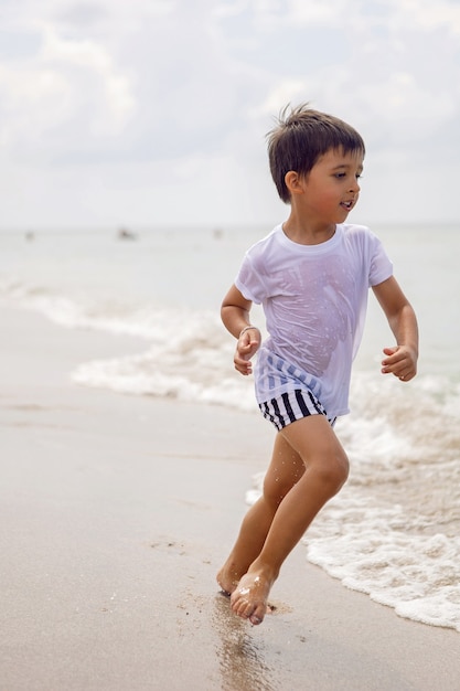 Niño en pantalones cortos a rayas y una camiseta blanca camina en una playa de arena y gafas de sol