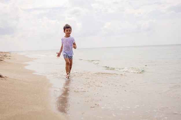 Niño en pantalones cortos a rayas y una camiseta blanca camina en una playa de arena y gafas de sol