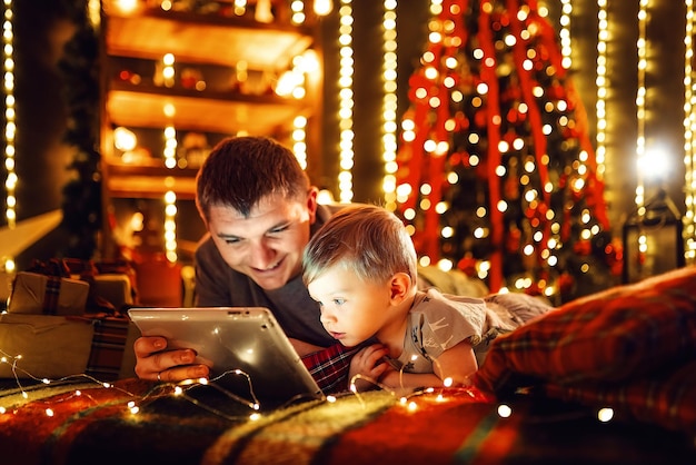 Niño con papá usando tablet pc en casa cerca del árbol de Navidad en una acogedora sala de estar decorada