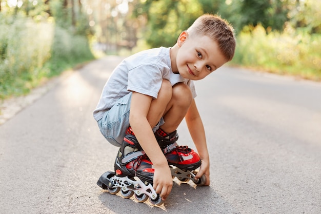 Niño en patinaje sobre ruedas sentado en la carretera, mirando a la cámara con una sonrisa, vestido con camiseta blanca y corta, el niño se pone en cuclillas mientras descansa patinar.