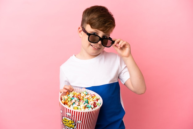 Niño pelirrojo aislado en la pared rosa con gafas 3d y sosteniendo un gran cubo de palomitas de maíz